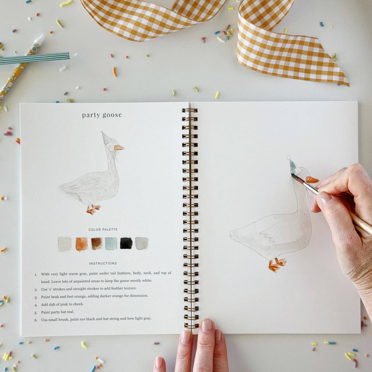 Open watercolor workbook with party goose drawing, spiral binding, and hand painting.