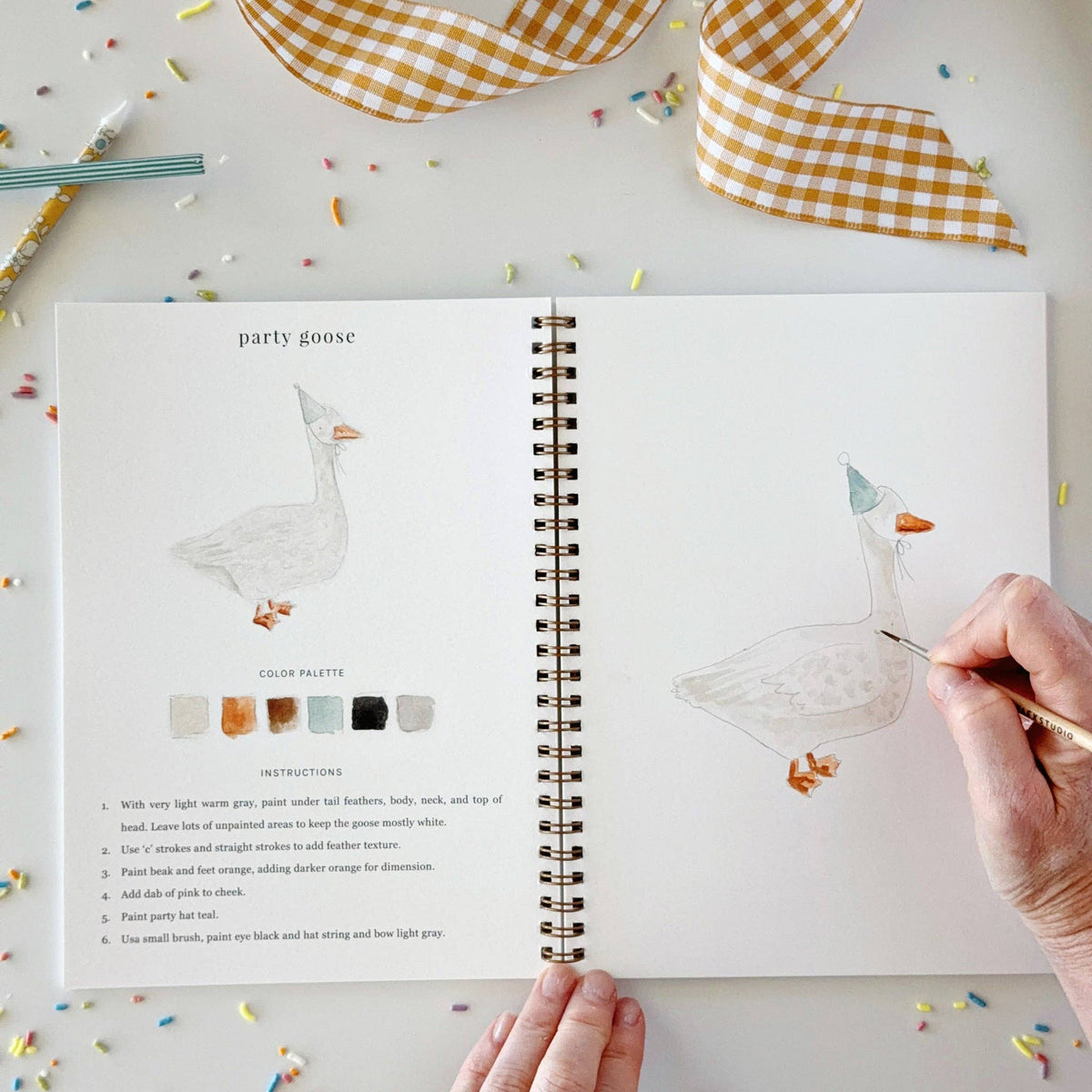 Open watercolor workbook showing party goose, spiral binding, and hand painting with brush.
