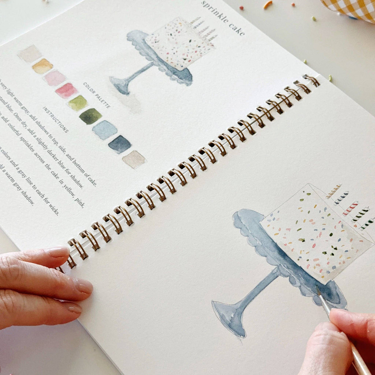 Watercolor workbook page with sprinkle cake illustration, spiral binding, and hand holding brush.