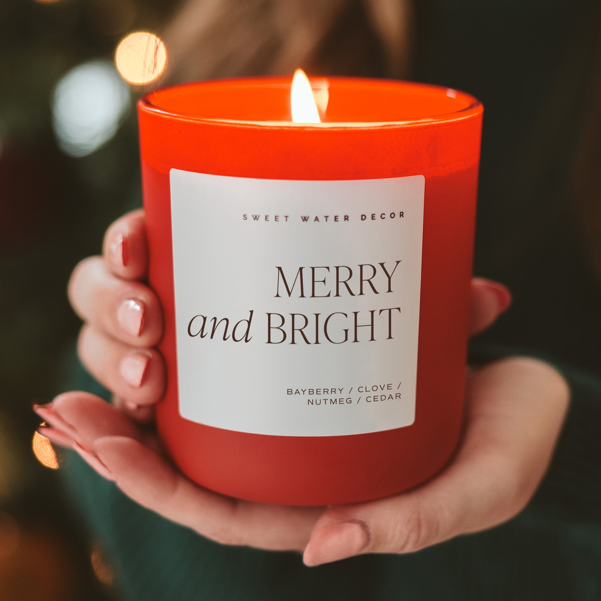 Red soy candle held in hands with “MERRY and BRIGHT” label and lit wick