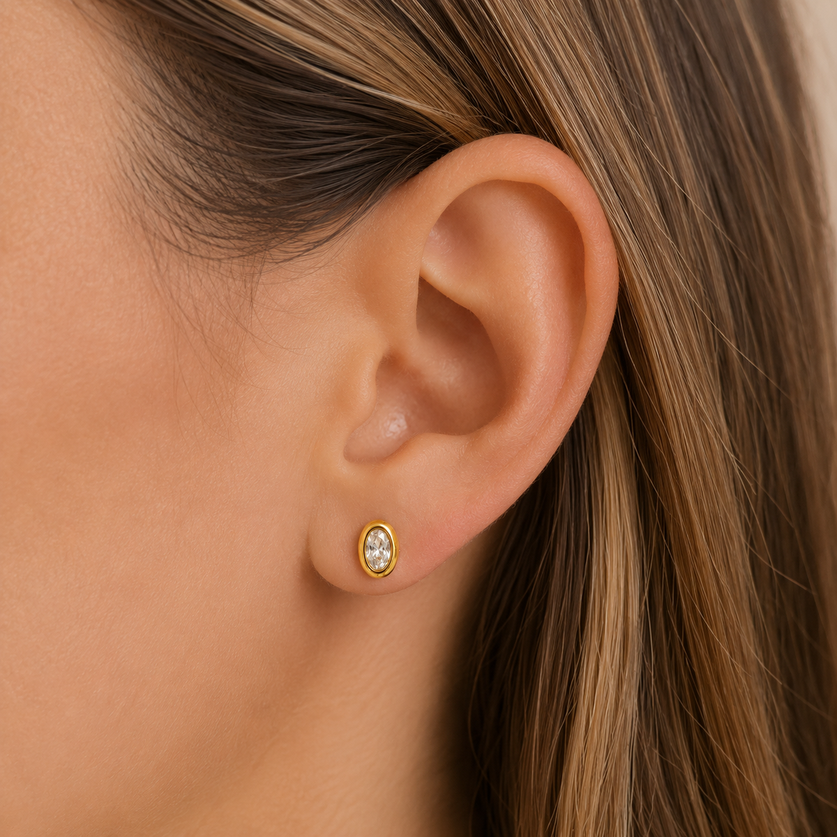 Oval CZ stud earring on ear, gold-tone bezel with clear center stone