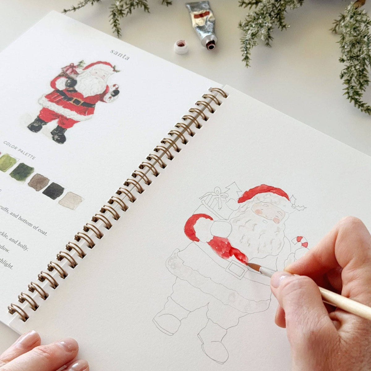 Christmas watercolor workbook open page with outlined Santa and hand painting with a small brush