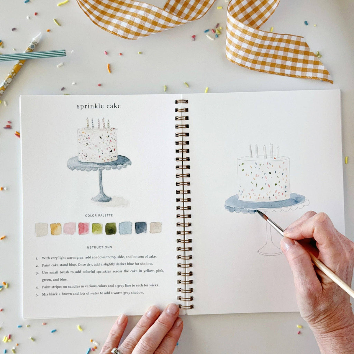 Open watercolor workbook with cake and palette, spiral binding, and hand painting page.