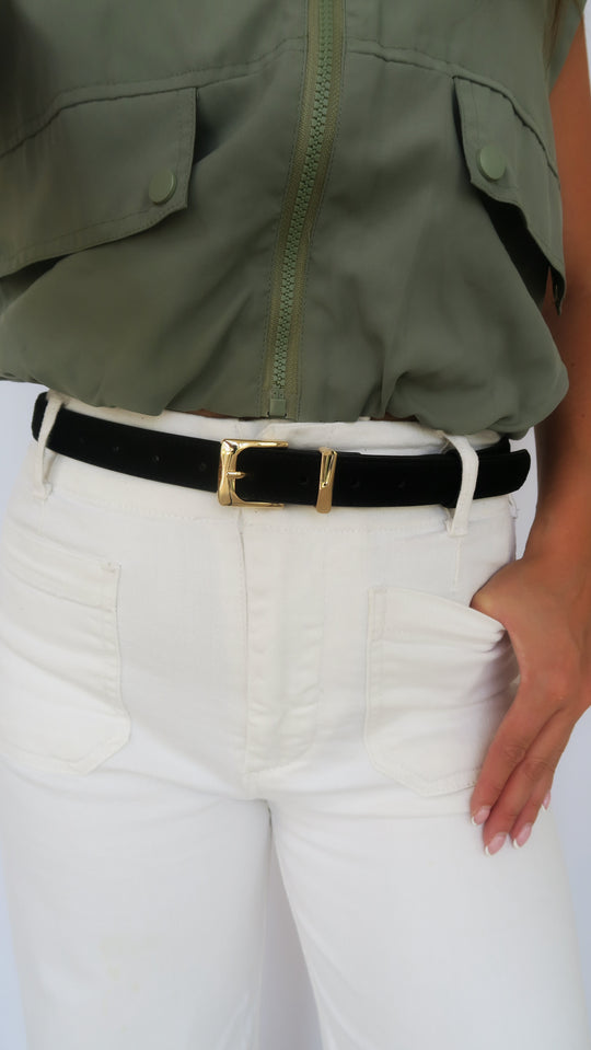 Black suede belt with gold-tone rectangular buckle worn on white pants, close-up.